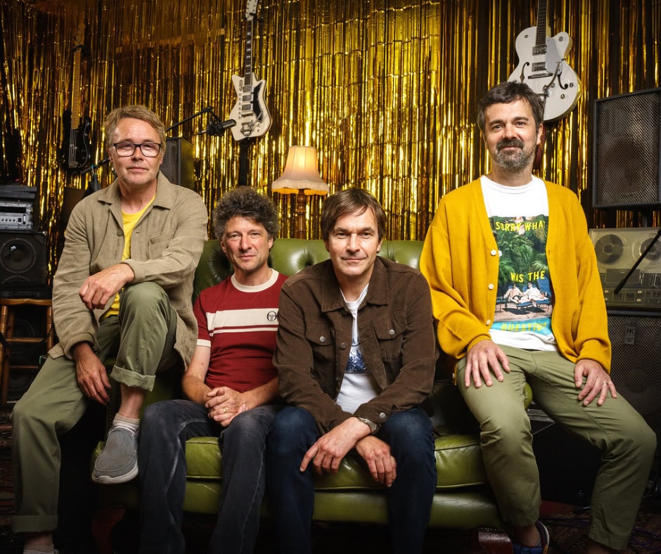 The Bluetones band sitting on a green sofa with musical instruments and gold curtain backdrop.